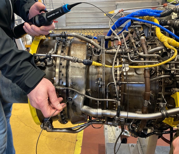 Inspector’s hands holding the IPLEX TX II borescope in one hand and guiding the insertion tube into an access point in a GE CT7 turbine engine for a helicopter