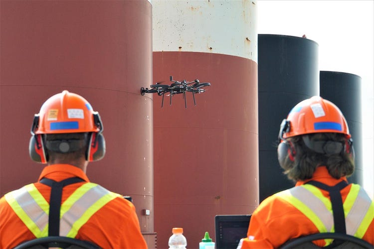 Dron siendo controlado por dos trabajadores a partir de un carro de seguridad. El dron permite medir el espesor de pared de un tanque de almacenamiento en una refinería de petróleo.